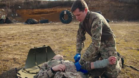 A-confident-man-in-camouflage-clothing-with-blue-elastic-medical-gloves-binds-the-hand-of-an-injured-man-with-a-tourniquet-during-combat-operations-in-the-steppe.-Providing-personal-assistance-to-wounded-soldiers-during-combat-operations