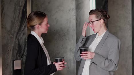 A-middle-aged-blonde-girl-with-glasses-in-a-business-suit-with-coffee-in-her-hands-communicates-with-her-businesswoman-colleague-a-young-blonde-girl-in-a-modern-office.-Dialogue-between-two-businesswoman-colleagues-in-the-office