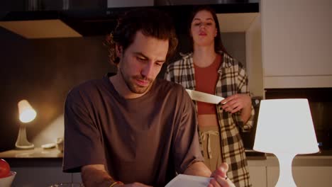 Scene-from-a-scary-movie:-a-brunette-guy-in-a-gray-T-shirt-reads-a-book-while-his-brunette-girlfriend-in-a-checkered-shirt-plays-with-a-knife-behind-him-in-a-modern-apartment-in-the-kitchen.-Staged-video