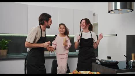 Happy-brunette-man-with-stubble-in-a-black-apron-together-with-his-brunette-wife-in-a-white-T-shirt-and-little-daughter-in-pink-clothes-dance-and-jump-while-in-the-kitchen-while-preparing-breakfast-on-a-happy-morning-in-a-modern-kitchen