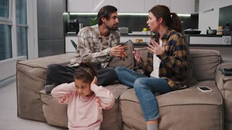 Sad-and-withdrawn-little-brunette-girl-in-a-pink-suit-covers-her-ears-with-her-palms-while-her-parents-in-a-checkered-brunette-shirt-are-arguing-while-sitting-on-the-sofa-in-a-modern-studio-apartment