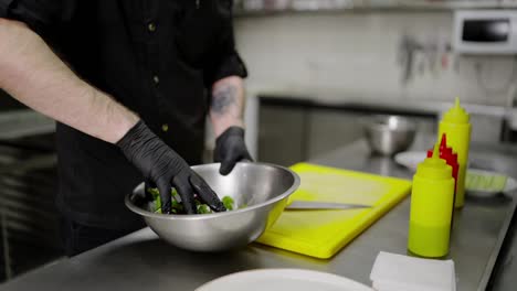 Close-up-a-confident-Chef-in-a-black-uniform-wearing-black-protective-rubber-gloves-mixes-a-salad-in-an-iron-bowl-before-presenting-and-serving-a-dish-in-a-restaurant-in-the-kitchen