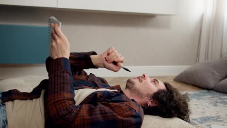 A-thoughtful-guy-comes-with-curly-hair-lying-on-a-pillow-and-writing-down-his-ideas-in-a-notepad-while-lying-on-the-floor-in-a-modern-apartment