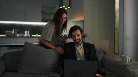 A-beautiful-brunette-girl-in-a-beige-T-shirt-is-interested-in-how-his-husbands-work-is-going-remotely.-A-brunette-with-stubble-in-a-black-jacket-and-beige-shorts-sits-on-a-gray-sofa-in-a-modern-studio-apartment-in-the-evening