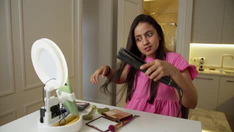 A-happy-Indian-girl-in-a-pink-dress-runs-her-Beauty-Blog-and-explains-in-a-video-how-to-properly-straighten-her-hair-using-a-special-straightening-iron-in-a-modern-apartment-online