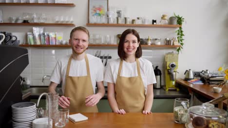 Portrait-of-a-happy-brunette-waiter-girl-in-a-yellow-apron-together-with-her-blond-male-colleague-with-a-beard-at-the-cash-register-in-a-cafe