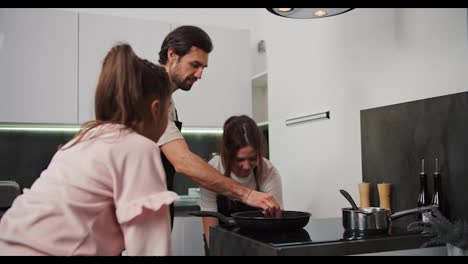 Una-Chica-Morena-Feliz-Con-Un-Delantal-Negro-Prepara-El-Desayuno-Con-Su-Novio-Y-Su-Pequeña-Hija-Por-La-Mañana-En-Una-Cocina-Moderna.-Una-Familia-Feliz-Preparando-El-Desayuno-Por-La-Mañana-En-La-Cocina-De-Un-Apartamento-Moderno.