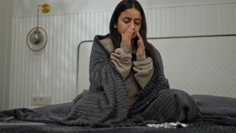 A-brunette-girl-is-sick-in-a-brown-sweater-and-a-dark-gray-blanket-wrapped-in-a-blanket-and-chooses-which-pills-to-take-while-coughing-and-experiencing-a-runny-nose-during-her-illness-in-a-modern-apartment-in-the-bedroom