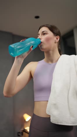 Vertical-video-of-a-confident-athletic-brunette-girl-in-a-purple-top-drinks-water-from-a-blue-and-sports-bottle-after-her-exercise-session-in-a-modern-apartment-at-home