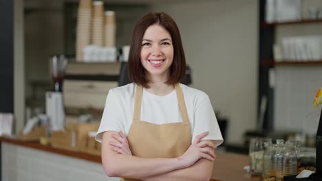 Portrait-of-a-happy-brunette-waiter-girl-in-a-white-T-shirt-and-a-light-brown-apron-posing-near-the-counter-in-a-cafe