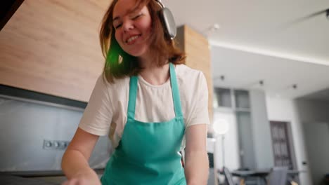 Happy-cheerful-brunette-cleaning-lady-girl-in-wireless-headphones-and-a-white-T-shirt-with-a-blue-front-washes-the-stove-in-the-kitchen-and-dances-while-having-fun-in-a-modern-apartment