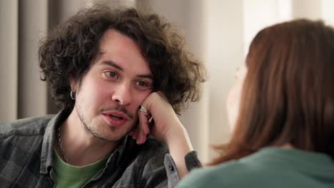 Over-the-shoulder,-a-happy-brunette-guy-with-curly-hair-and-a-mustache-props-his-head-with-his-hand-and-talks-to-a-brunette-girl-at-home-on-the-sofa