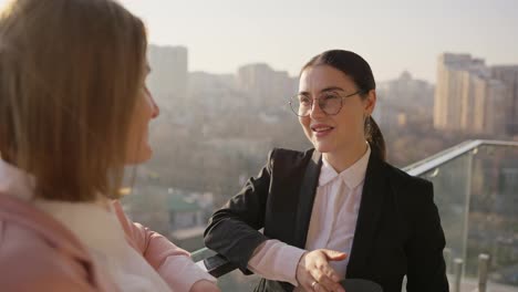 Over-the-shoulder-a-confident-brunette-businesswoman-girl-in-round-glasses-and-a-black-uniform-communicates-with-her-colleague-while-standing-on-the-terrace-in-a-modern-office-overlooking-the-city-sunny-weather