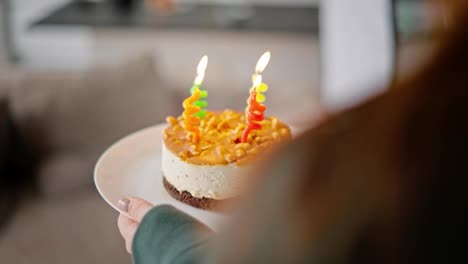 Por-Encima-Del-Hombro,-Una-Chica-Morena-Con-Una-Chaqueta-Verde-Trae-Un-Pequeño-Pastel-Con-Velas-Para-Su-Papá-En-Honor-A-Su-Cumpleaños.-Un-Anciano-Feliz-Con-Cabello-Gris-Y-Una-Barba-Exuberante-Con-Una-Camisa-Rosa-Apaga-Las-Velas-Y-Agradece-A-Su-Hija-Adulta-Por-Sus-Deseos-De-Cumpleaños-En-Un-Apartamento-Moderno.