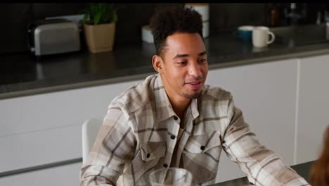 Close-up-of-a-happy-young-man-with-Black-skin-color-in-a-checkered-beige-shirt-drinks-orange-juice-with-his-girlfriend-during-his-breakfast-in-the-kitchen