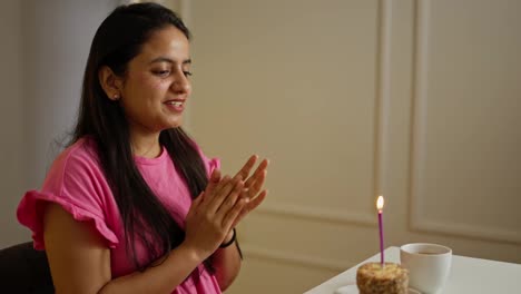 Side-view-of-a-happy-brunette-Indian-girl-in-a-pink-dress-blowing-out-a-candle-on-a-small-cake-and-clapping-her-hands-during-a-birthday-celebration-in-a-modern-apartment-during-the-day