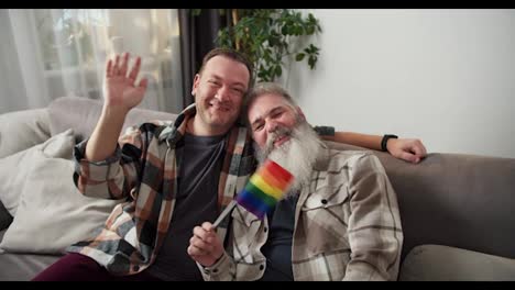 Retrato-De-Un-Hombre-Feliz-Con-Cabello-Gris-Y-Una-Exuberante-Barba-Blanca-Con-Una-Camisa-A-Cuadros-Que-Sostiene-Una-Bandera-Lgbt-Junto-Con-Su-Novio-Moreno-Con-Una-Camisa-A-Cuadros-Sentado-En-Un-Sofá-Gris-En-Un-Apartamento-Moderno-Por-La-Noche.