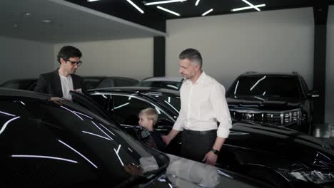 A-confident-brunette-guy-in-a-business-suit-an-assistant-at-a-car-dealership-communicates-with-a-middle-aged-man-and-lets-his-little-son-get-into-the-car