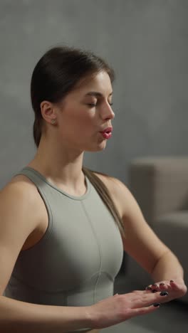 Vertical-video-of-a-confident-brunette-girl-in-a-gray-swamp-doing-yoga-and-breathing-practice-in-a-modern-apartment-at-home.-A-confident-athletic-brunette-girl-in-a-gray-swamp-is-engaged-in-breathing-practices-to-develop-her-spiritual-peace