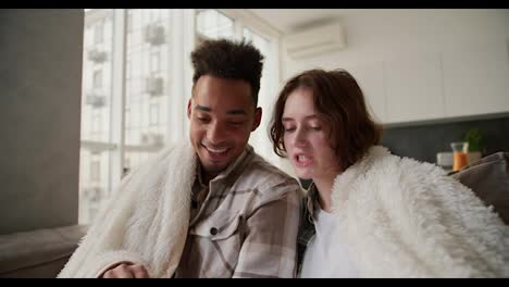 Close-up-a-happy-young-man-with-Black-skin-a-brunette-with-stubble-in-a-checkered-cream-shirt-sits-on-a-modern-sofa-with-his-young-girlfriend-wrapped-in-a-white-woolen-blanket.-They-read-funny-and-happy-stories-to-each-other-in-a-modern-apartment