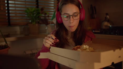 Confident-brunette-girl-in-red-wireless-headphones-with-glasses-studying-foreign-languages-and-learning-new-things-while-making-a-snack-and-eating-pizza-from-a-packaging-box-in-a-modern-apartment-in-the-kitchen