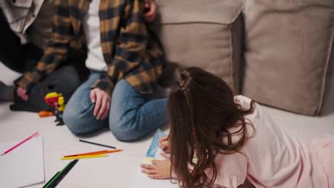 Close-up,-a-little-brunette-girl-in-pink-clothes-draws-lying-on-the-floor-her-parents-look-at-her-a-brunette-man-with-stubble-in-a-checkered-shirt-and-his-brunette-wife-in-a-white-T-shirt-and-checkered-shirt-near-a-gray-and-brown-sofa-in-a-modern-studio-apartment