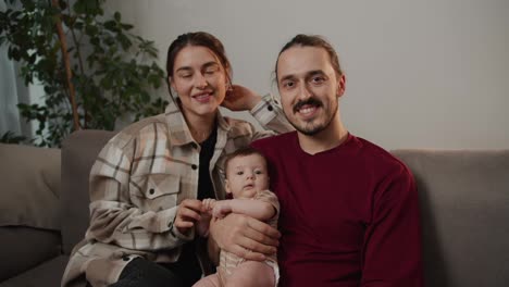 Porträt-Einer-Glücklichen-Jungen-Familie,-Ein-Brünettes-Mädchen-In-Einem-Karierten-Hemd-Zusammen-Mit-Ihrem-Brünetten-Ehemann-In-Einem-Roten-T-Shirt-Halten-Ihre-Kleine-Tochter-In-Den-Armen-Und-Sitzen-Auf-Einem-Grauen-Sofa-In-Einer-Modernen-Wohnung