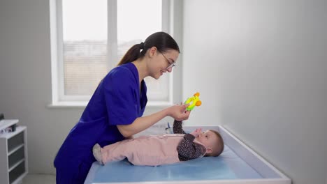 Happy-brunette-girl-with-glasses-in-a-blue-doctors-uniform-plays-with-a-little-girl-baby-during-an-appointment-with-pediatricians-in-a-modern-clinic