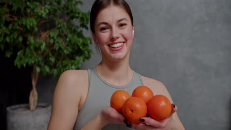 Portrait-of-a-confident-and-happy-young-brunette-girl-in-a-gray-sports-top-who-holds-four-orange-oranges-in-her-hands-near-a-large-indoor-plant-in-a-modern-apartment-at-home