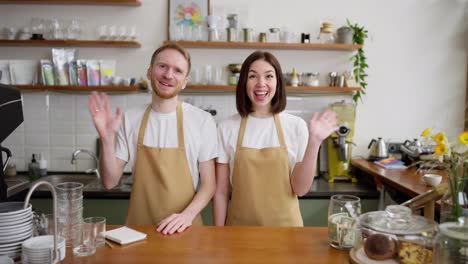 Portrait-Happy-waiters-and-barista-blond-man-and-brunette-girl-wave-and-welcome-everyone-to-their-cafe