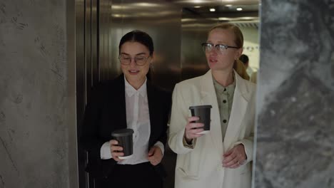 Two-confident-businesswomen-in-black-and-white-uniforms-and-glasses-come-out-of-the-elevator-while-starting-their-working-day-drink-coffee-and-chat-while-walking-around-a-modern-office