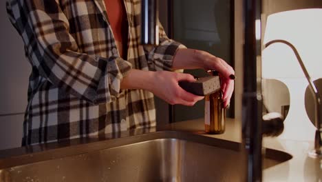 A-confident-brunette-girl-in-a-checkered-shirt-takes-out-a-gray-dishwashing-sponge-pours-a-special-detergent-on-it-and-washes-the-dishes-in-the-evening-during-her-routine-in-a-modern-apartment