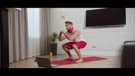 Happy-confident-blond-athletic-man-with-stubble-in-a-white-T-shirt-and-red-shorts-doing-squats-during-his-morning-exercises-while-watching-a-guide-on-his-laptop-in-a-modern-bright-apartment-at-home