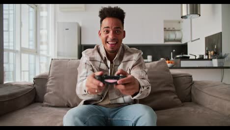 Portrait-of-a-happy-young-Black-skinned-brunette-man-with-stubble-who-sits-on-a-modern-sofa-and-plays-a-video-game-and-rejoices-at-his-victory-using-a-black-joystick-in-a-modern-studio-apartment