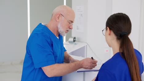 A-confident-male-doctor-of-advanced-age-with-a-gray-beard-in-glasses-and-in-a-blue-uniform-communicates-with-his-colleague-a-brunette-girl-at-the-reception-in-a-modern-clinic-during-the-working-day