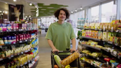 A-brunette-guy-with-curly-hair-walks-with-a-cart-filled-with-groceries-and-looks-at-the-goods-in-a-grocery-supermarket