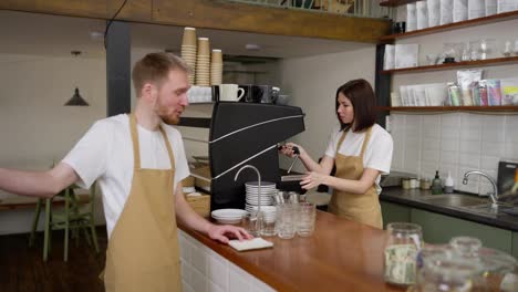 A-confident-brunette-barista-girl-in-a-yellow-apron-communicates-with-a-guy-waiter-and-prepares-his-order-while-working-in-a-cafe