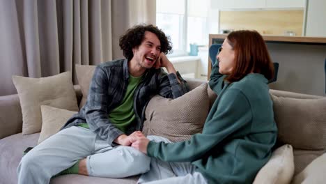 Happy-guy-with-curly-hair-laughs-loudly-and-has-fun-while-communicating-with-his-brunette-girlfriend-while-sitting-on-the-sofa-at-home-in-a-modern-apartment