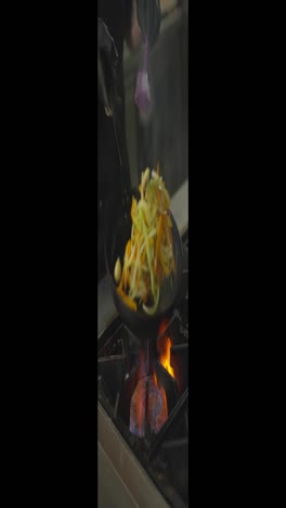 Close-up-vertical-video-of-a-confident-chef-in-black-gloves-frying-vegetables-on-a-gas-burner-in-the-kitchen-of-a-restaurant.-And-the-cook-fries-vegetables-in-a-frying-pan-turning-them-over-and-shaking-the-pan-masterfully-managing-the-vegetables