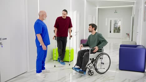 A-confident-male-doctor-with-a-gray-beard-and-glasses-in-a-blue-uniform-communicates-with-two-guys-after-injuries-during-rehabilitation-with-crutches-and-a-special-chair-for-the-disabled-in-a-modern-clinic