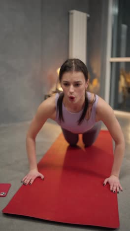 Close-up-vertical-video-of-a-confident-brunette-girl-in-a-sports-summer-uniform-doing-push-ups-and-sprints-on-a-red-sports-mat-in-a-sports-mat-in-a-modern-apartment-at-home