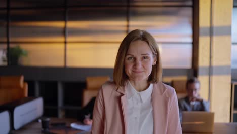 Portrait-of-a-confident-middle-aged-blonde-businesswoman-in-a-pink-business-suit-near-her-colleagues-at-her-desk-in-a-sunny-office