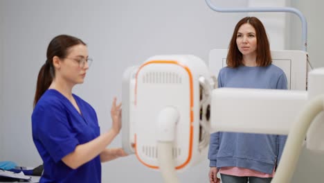 A-confident-brunette-girl-in-round-glasses.-A-doctor-in-a-blue-uniform-does-fluorography-for-a-brunette-girl-in-a-jacket-in-a-modern-clinic