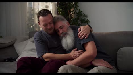 A-middle-aged-brunette-man-with-stubble-in-a-gray-T-shirt-hugs-his-sad-boyfriend-a-man-with-gray-hair-color-and-a-lush-gray-beard-while-sitting-on-a-sofa-in-a-modern-apartment-in-the-evening.-Showing-love-and-care-in-difficult-times-to-a-couple-of-middle-aged-LGBT-men-in-a-modern-apartment