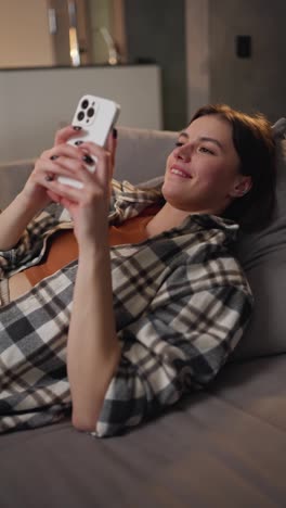 Vertical-video-of-a-happy-and-joyful-brunette-girl-in-a-checkered-shirt-and-an-orange-top-looks-at-the-social-network-using-a-white-smartphone-and-communicates-online-in-a-modern-apartment-lying-on-the-sofa