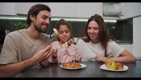 Un-Hombre-Moreno-Feliz-Con-Barba-Incipiente-Con-Una-Camiseta-Beige-Junto-Con-Su-Esposa-Morena-Con-Una-Camiseta-Blanca-Y-Una-Pequeña-Hija-Con-Ropa-Rosa-Están-Desayunando-En-Un-Apartamento-Moderno-En-Una-Mesa-De-Comedor-Negra-Por-La-Mañana.