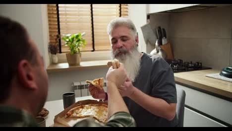 Over-the-shoulder-a-brunette-man-in-a-plaid-shirt-feeds-his-middle-aged-man-with-gray-hair-color-and-a-full-gray-beard-during-their-pizza-lunch-in-the-kitchen-in-a-modern-apartment