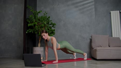 Confident-brunette-girl-in-a-gray-top-sports-uniform-and-green-pants-does-yoga-and-stretching-on-a-red-sports-mat-in-a-modern-apartment-near-a-large-indoor-plant-using-online-lessons-via-a-laptop