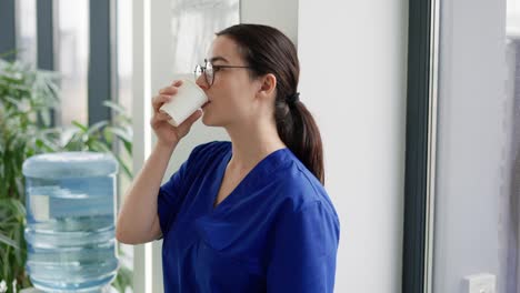 A-tired-brunette-girl-in-round-glasses-a-doctor-in-a-blue-uniform-drinks-water-from-a-white-plastic-cup-in-a-modern-clinic-during-her-break