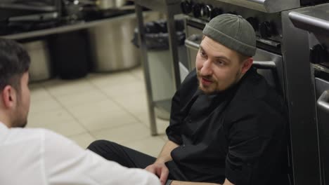 Comunicación-Entre-Un-Cocinero-Profesional-Con-Uniforme-Negro-Y-Un-Camarero-Durante-Un-Breve-Descanso-En-La-Cocina-Mientras-Están-Sentados-En-El-Suelo-Apoyados-En-La-Mesa-Durante-Un-Ajetreado-Día-De-Trabajo.